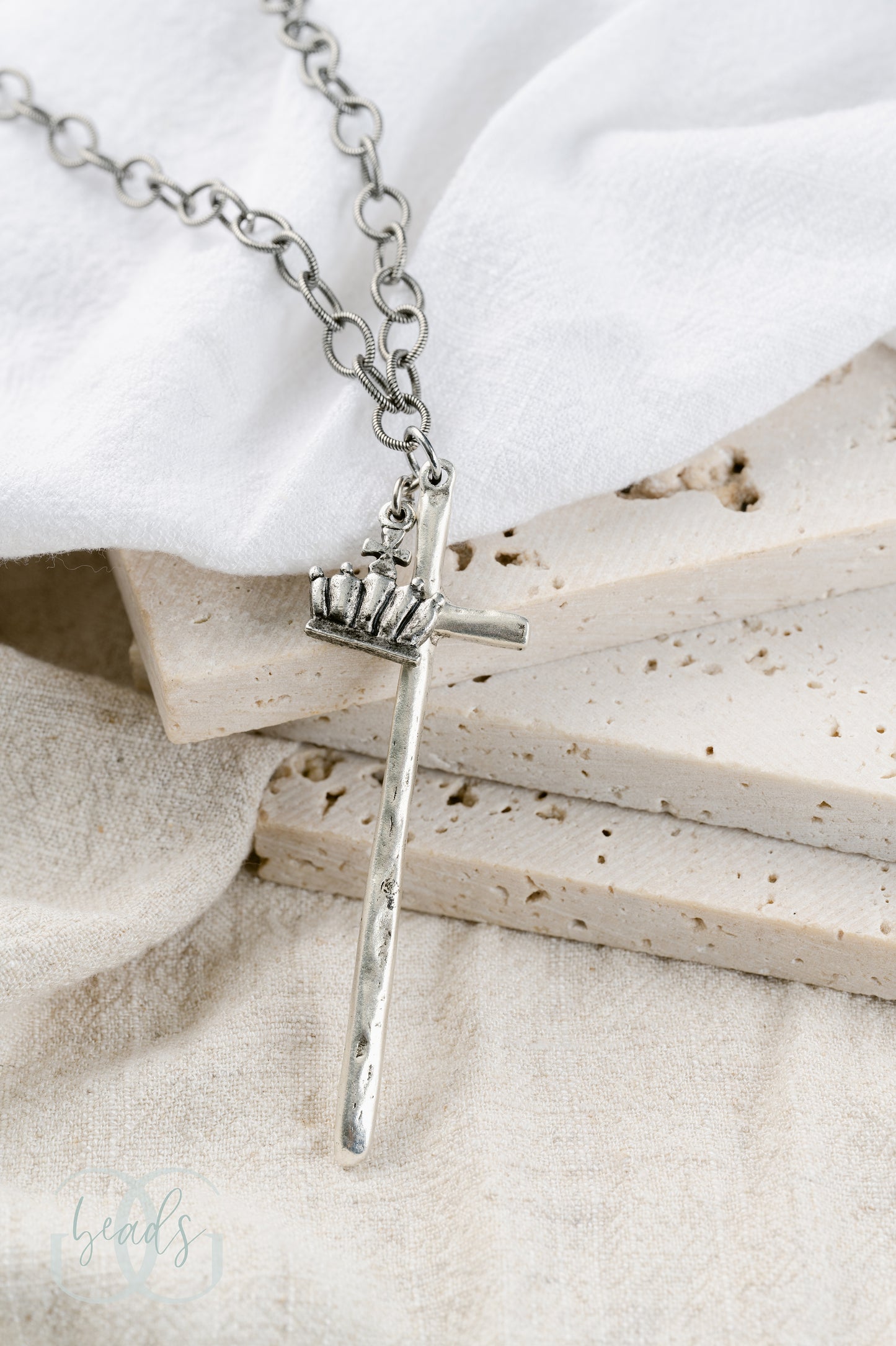 A silver necklace featuring a long, hammered cross with a crown on it.