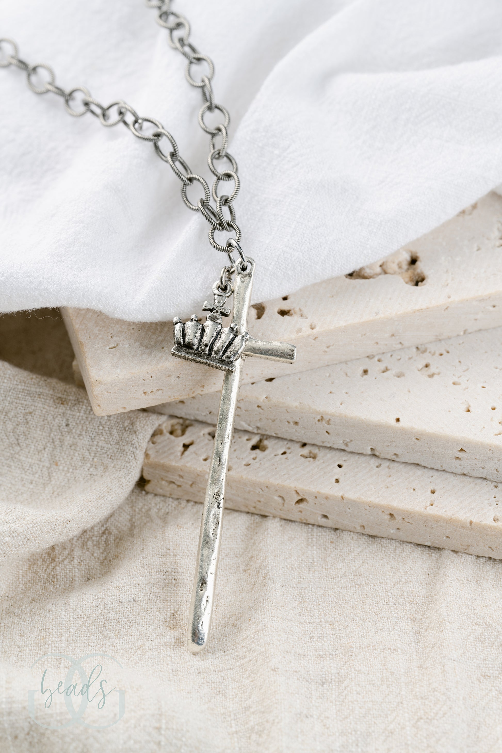 A silver necklace featuring a long, hammered cross with a crown on it.