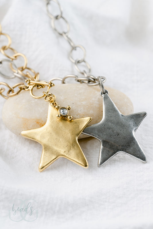 A chunky chain with a large star pendant, shown in silver and gold. The gold necklace has a front toggle closure and a crystal hanging from it. The silver has a standard clasp on the back.