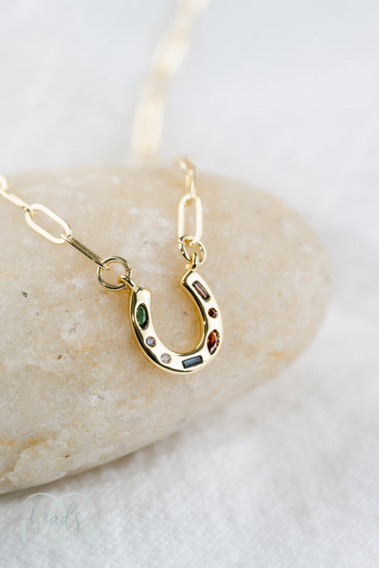 A gold horseshoe necklace on a dainty chain with seven crystals in it. The crystals are green, white, blue, and red.