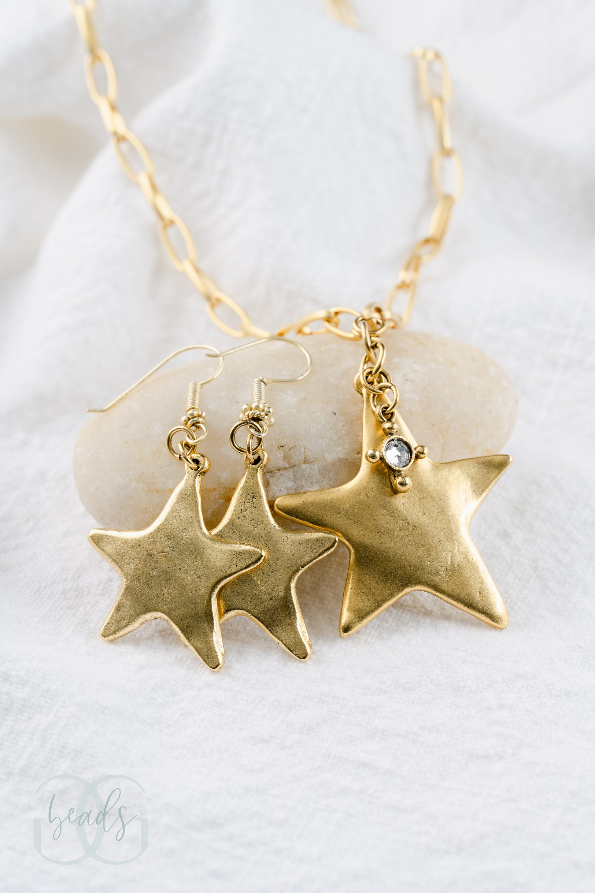 A gold chain featuring a large gold star pendant with a small crystal hanging from it. It is shown here with a matching pair of earrings - a gold star pendant.
