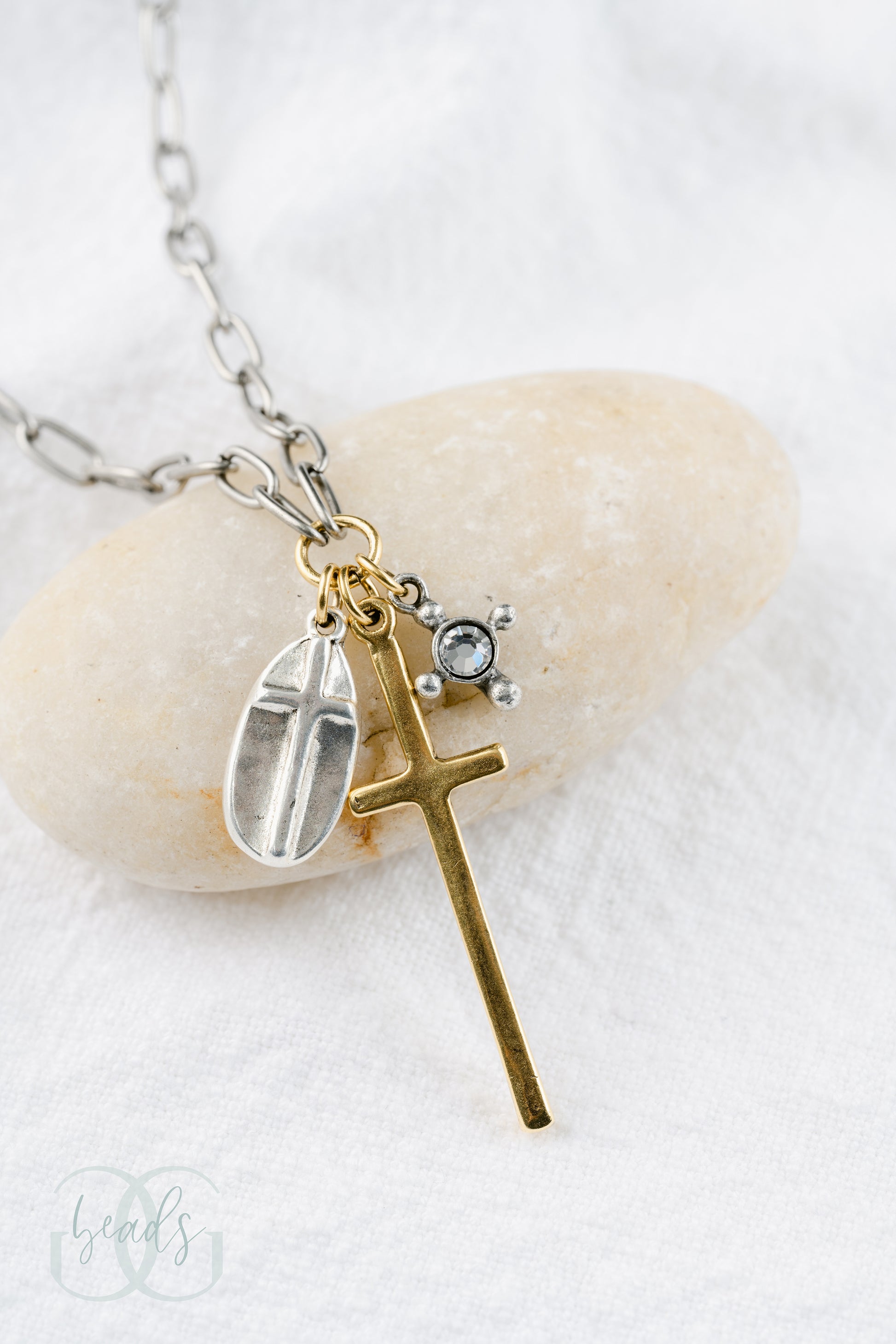 A silver chain featuring a triple cross design - one charm is a silver oval shape with a cross on top of it, the largest charm is a long gold cross, the smallest charm is a crystal with four dots around it symbolizing a cross.