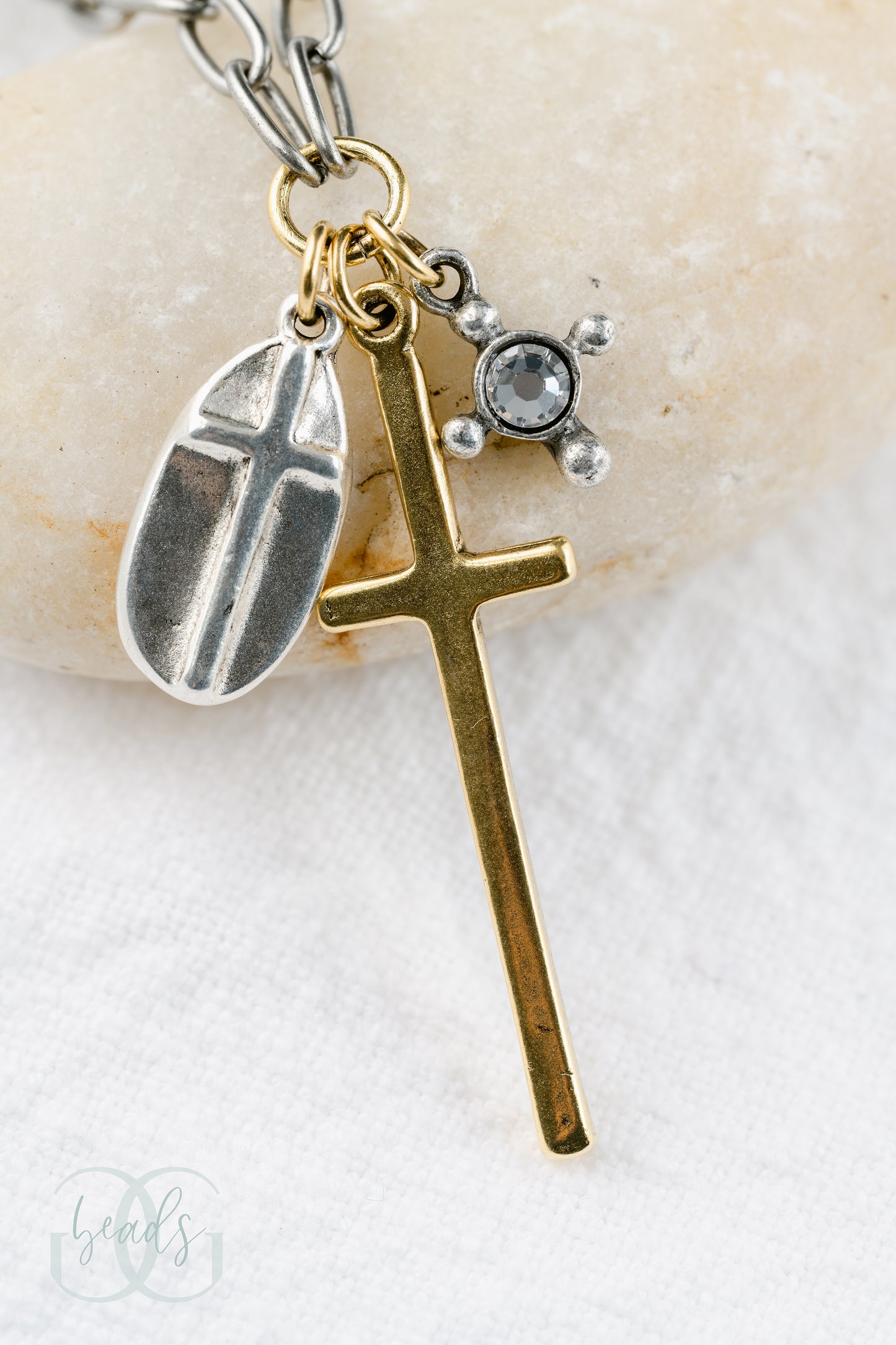 A silver chain featuring a triple cross design - one charm is a silver oval shape with a cross on top of it, the largest charm is a long gold cross, the smallest charm is a crystal with four dots around it symbolizing a cross.
