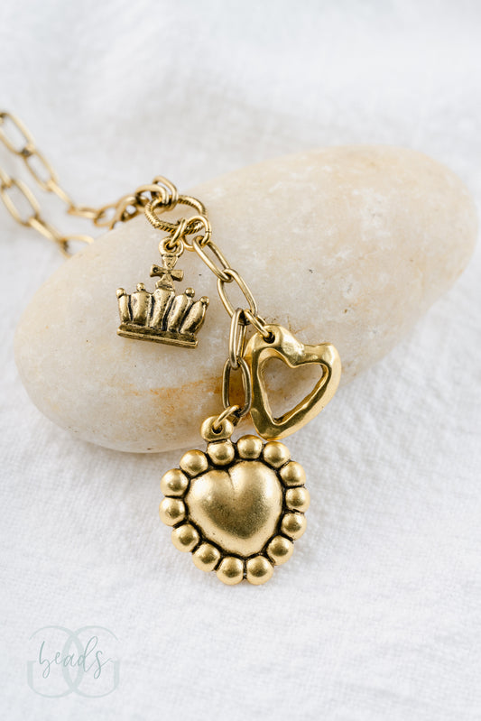 A gold lariat style necklace with an open heart charm, a crown charm and a puffy heart charm.