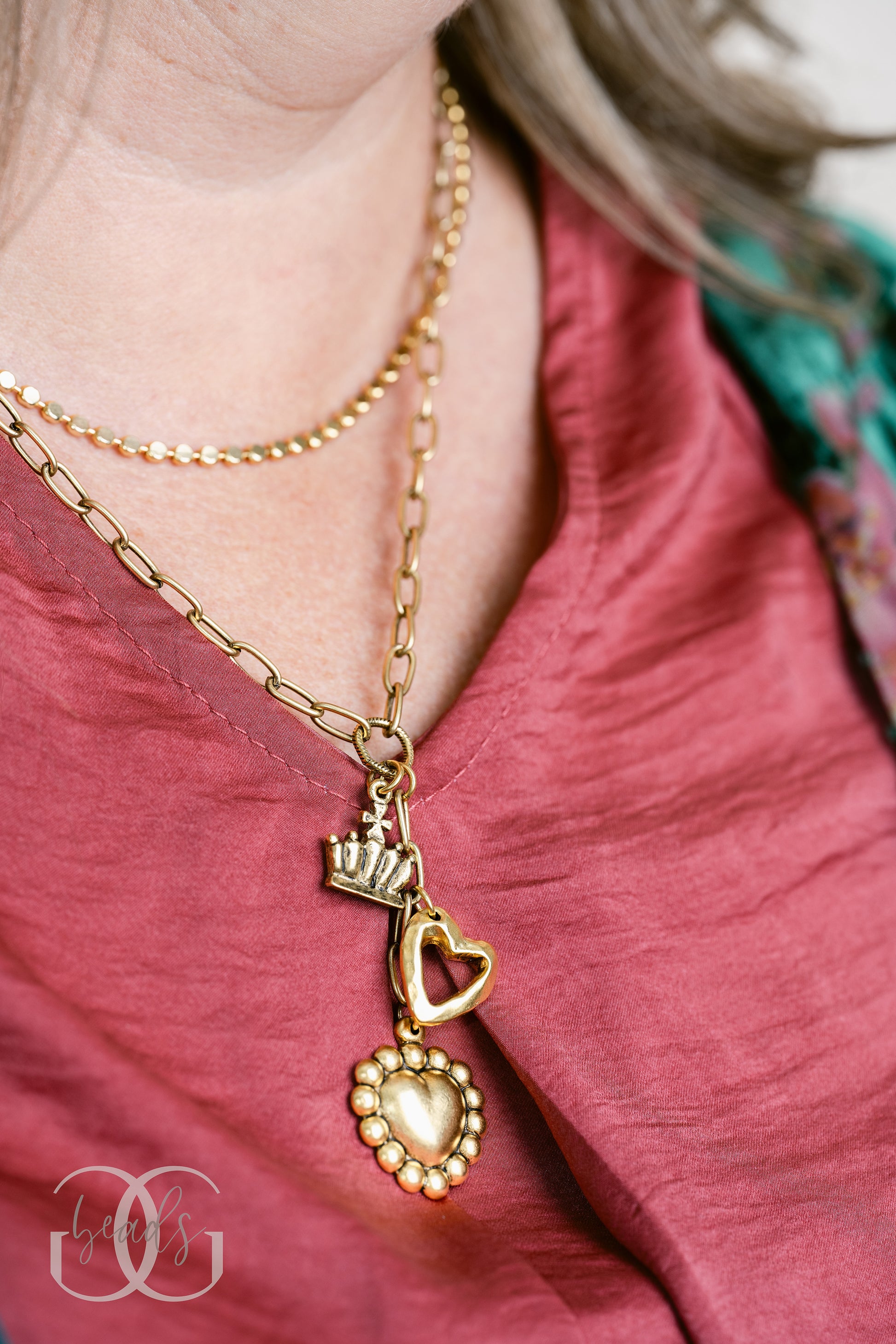 A gold lariat style necklace with an open heart charm, a crown charm and a puffy heart charm and being paired with our Dot Necklace.