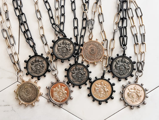 A picture of nine Mix it Up Necklaces. Each necklace is on a large paperclip chain, black or mixed metal, with a coin surrounded by a bezel. The coins are gold, silver, and black and the bezels are also gold, silver, and black.