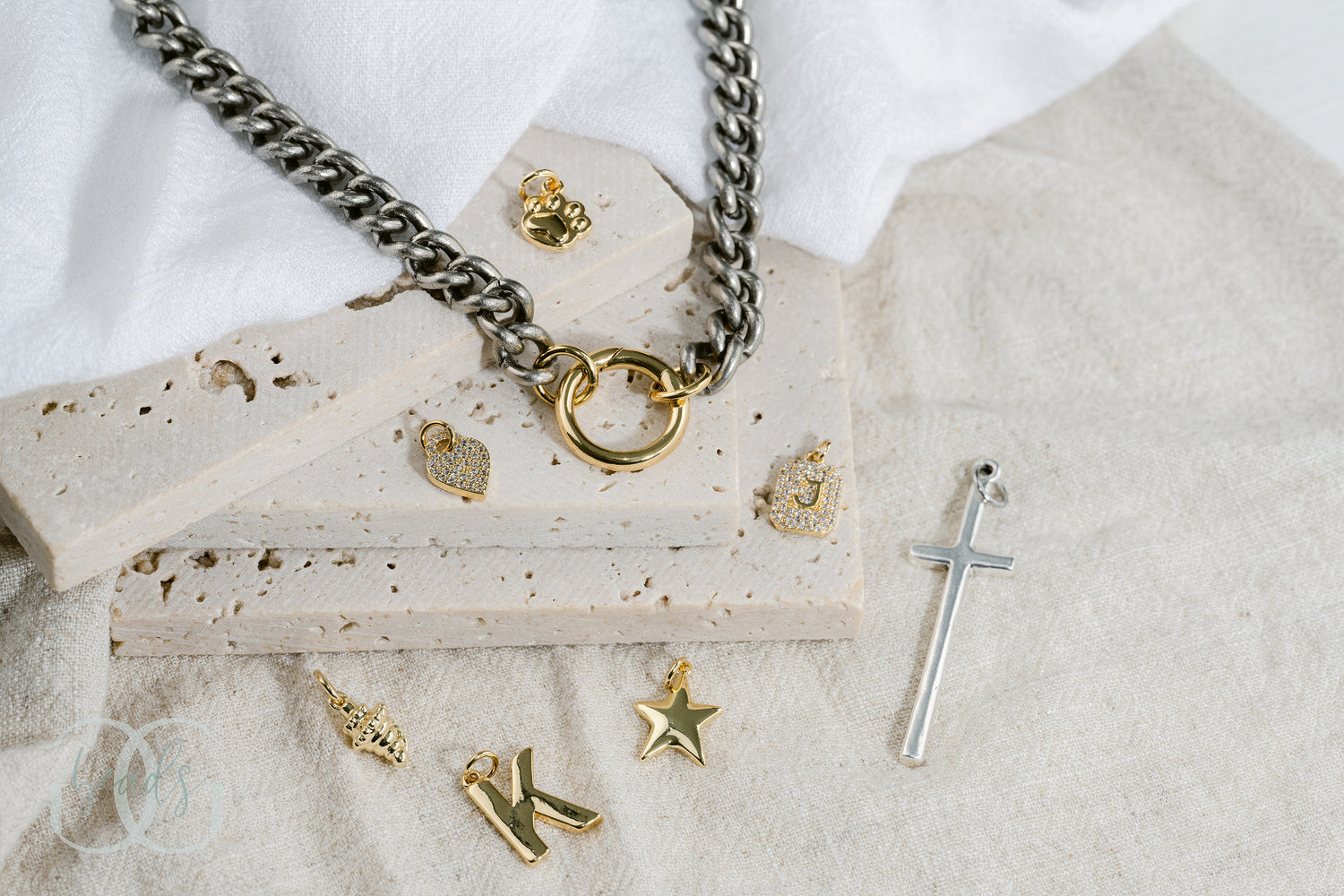 A chunky, silver chain link necklace with a gold spring gate front-closure clasp on it so you can add charms to it. Seven charms surround the necklace these charms are: a gold paw print, a gold pave crystal heart, a gold pave crystal initial, a gold seashell, a gold initial, a gold star, a silver cross.