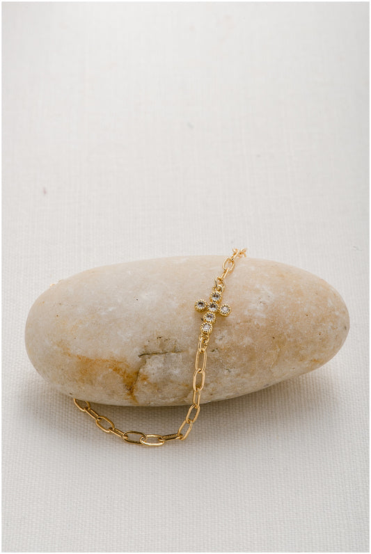 A gold paperclip chain with a round cut crystal cross charm embedded in it.