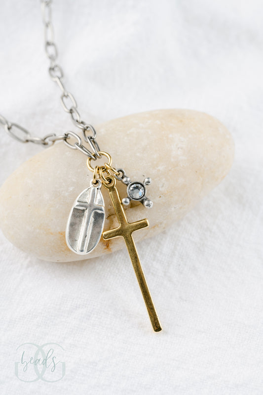 A silver chain featuring a triple cross design - one charm is a silver oval shape with a cross on top of it, the largest charm is a long gold cross, the smallest charm is a crystal with four dots around it symbolizing a cross.