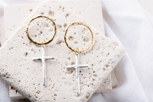 Round, gold earrings with a long, silver cross pendant. 